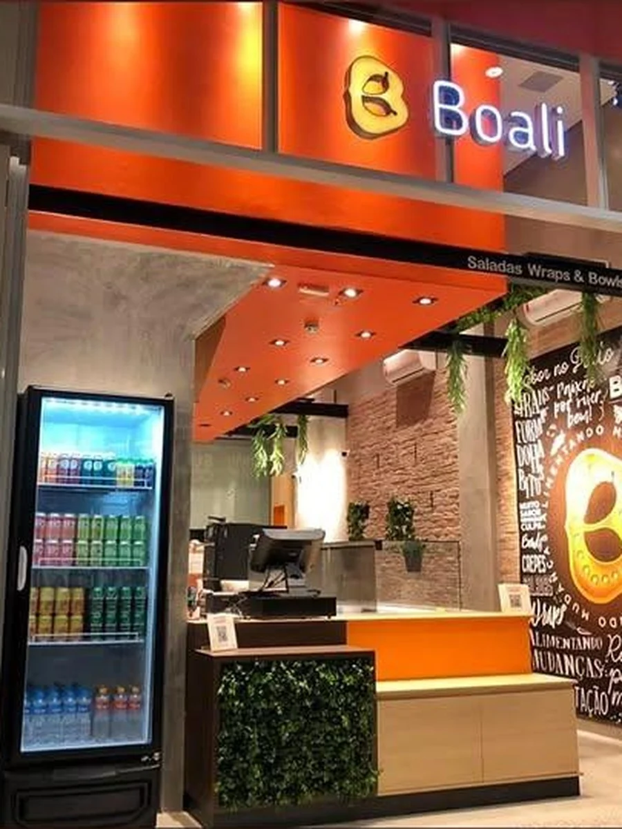 Vista frontal da loja à noite, com o painel decorativo e o expositor refrigerado visíveis pela vitrine.