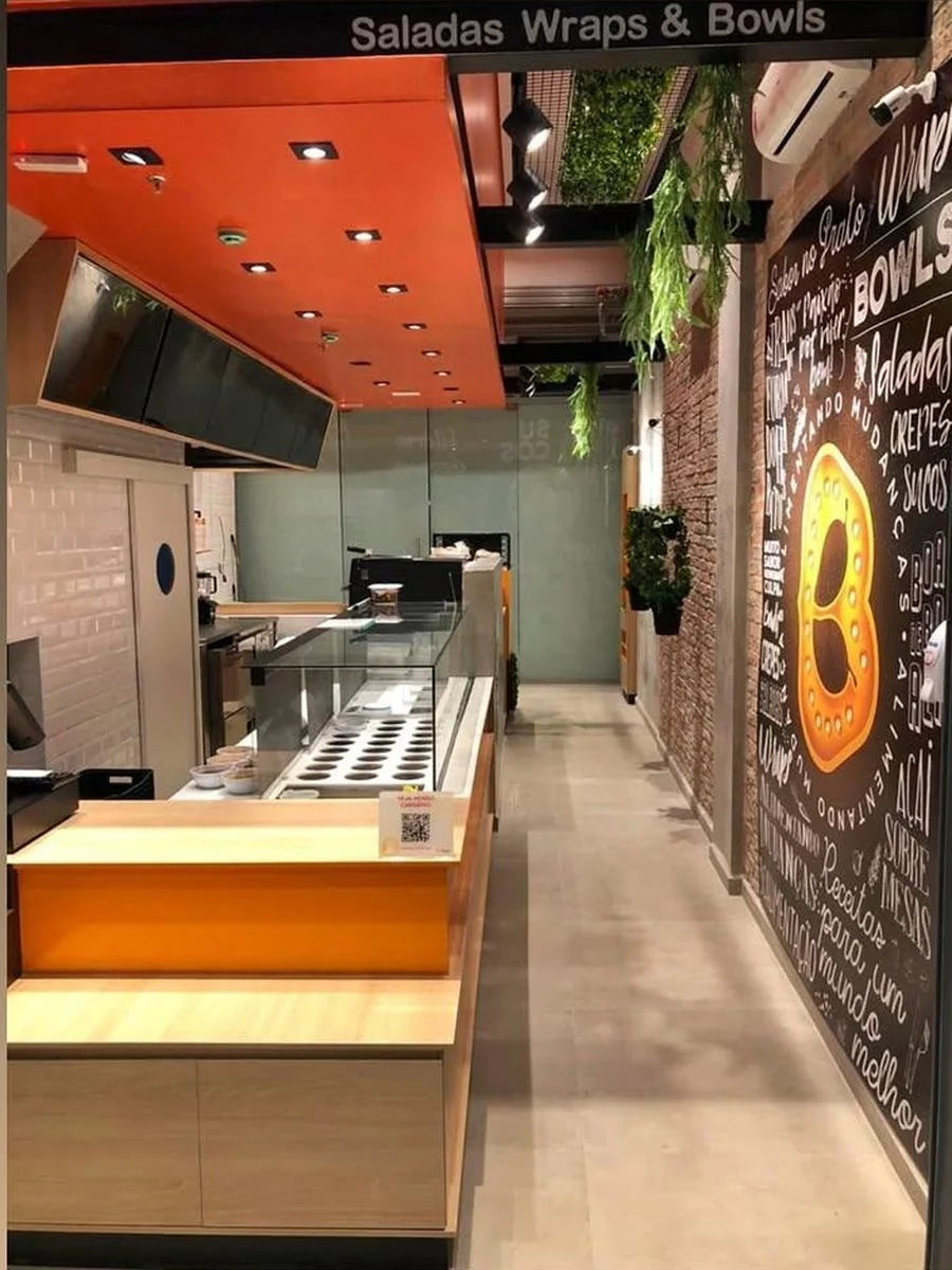 Interior da loja: linha de serviço com vitrine refrigerada, coifa e parede de tijolo com painel escrito à mão.